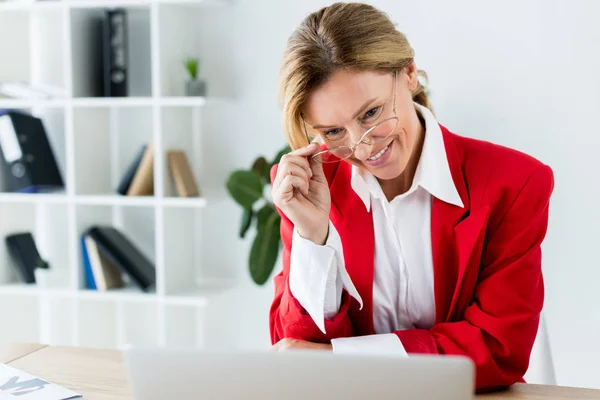 Donna d'affari attraente sorridente che tocca occhiali e guarda il computer portatile in ufficio — Foto stock