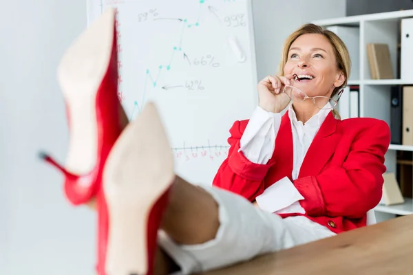 Attractive businesswoman laughing and sitting with legs on table in office — Stock Photo