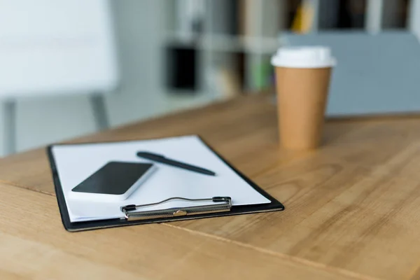 Smartphone and disposable coffee cup with clipboard on wooden table in office — Stock Photo
