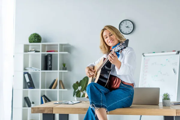 Smiling beautiful businesswoman playing acoustic guitar in office — Stock Photo
