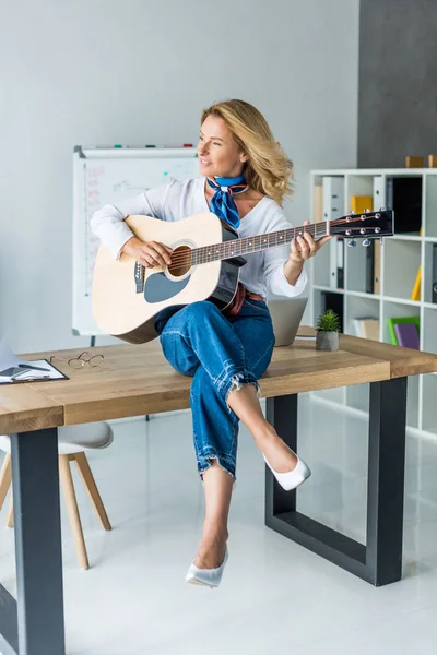 Smiling attractive businesswoman playing acoustic guitar in office — Stock Photo