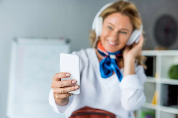 Feliz atractiva mujer de negocios en auriculares tomando selfie con teléfono inteligente en la oficina - foto de stock