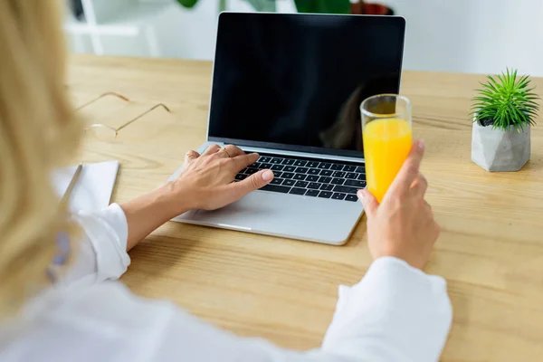 Image recadrée de femme d'affaires travaillant avec un ordinateur portable et tenant un verre de jus dans le bureau — Photo de stock