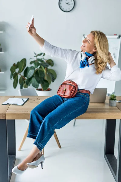 Attractive businesswoman sitting on table in office and taking selfie with smartphone — Stock Photo