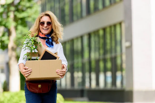 Sonriente atractiva mujer sosteniendo caja de papel con cosas de oficina en la calle - foto de stock