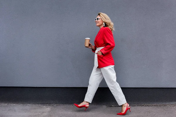 Vista lateral de mujer atractiva en chaqueta roja caminando con café en taza de papel en la calle - foto de stock
