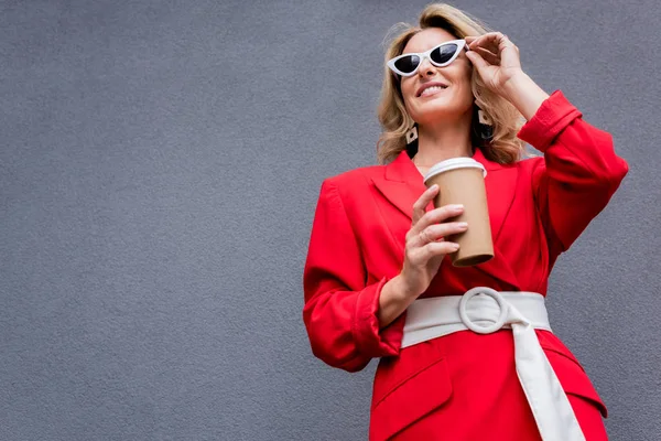Attractive woman in red jacket holding coffee in paper cup and touching sunglasses on street — Stock Photo