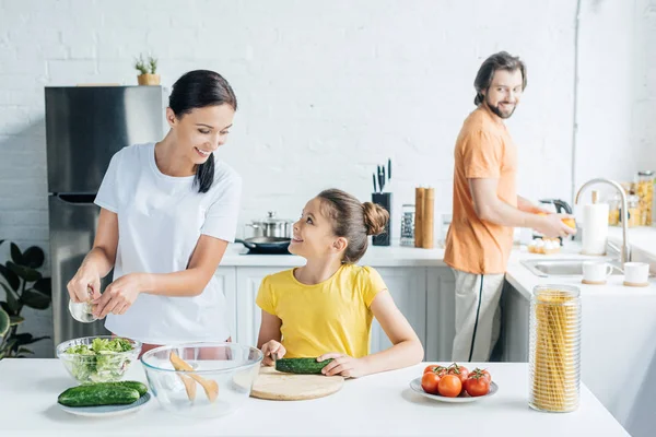 Mother and daughter preparing salad while father washing dishes on background at kitchen — Stock Photo