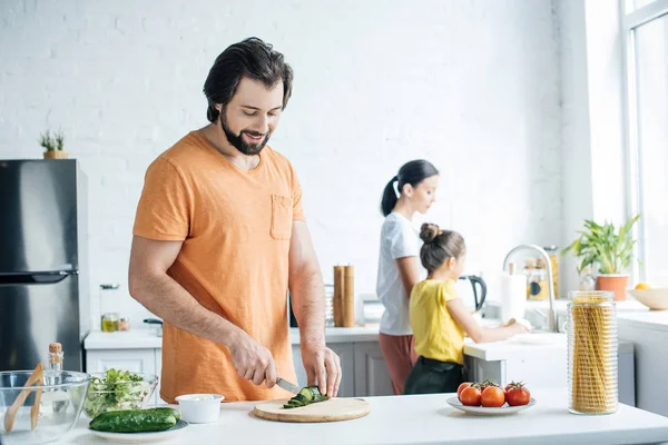 Felice giovane padre taglio cetriolo per insalata mentre la moglie e la figlia lavare i piatti in cucina — Foto stock