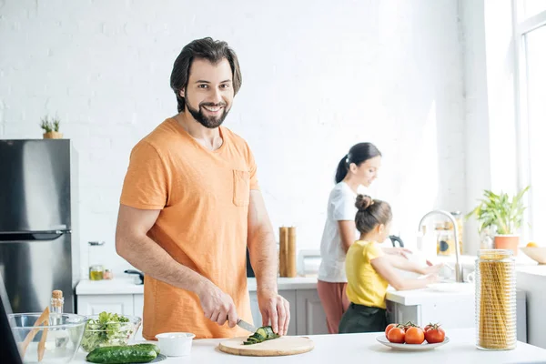 Lächelnder junger Vater schneidet Gurken für Salat, während seine Frau und Tochter in der Küche Geschirr spülen — Stockfoto