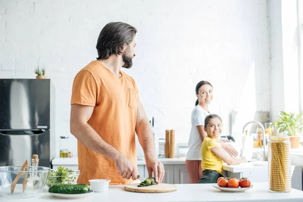 Bärtiger junger Vater schneidet Gurken für Salat, während seine Frau und Tochter in der Küche Geschirr spülen — Stockfoto