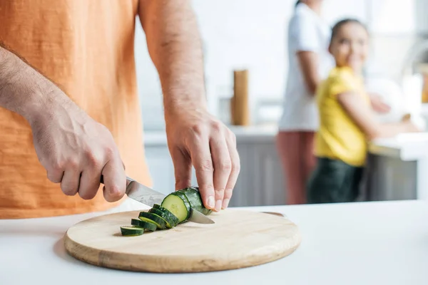 Recortado tiro de padre cortando pepino para ensalada mientras su esposa e hija lavar platos en la cocina - foto de stock