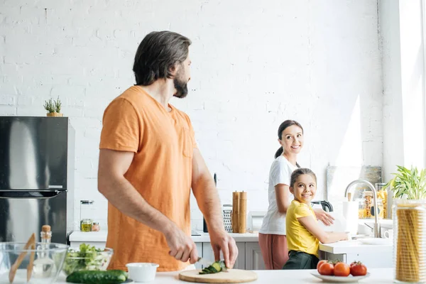 Attraktiver junger Vater schneidet Gurken für Salat, während seine Frau und Tochter in der Küche Geschirr spülen — Stockfoto