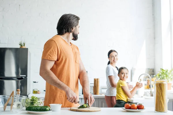 Schöner junger Vater schneidet Gurken für Salat, während seine Frau und Tochter in der Küche Geschirr spülen — Stockfoto