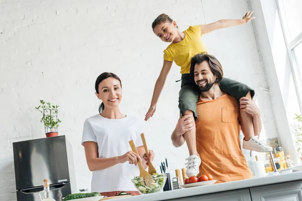 Jovem feliz preparando salada enquanto sua filha montando nos ombros do marido na cozinha — Fotografia de Stock
