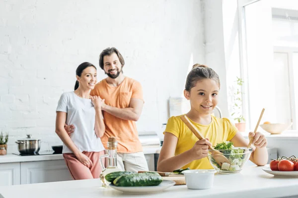 Sorrindo menina preparando salada enquanto seus pais de pé embaçado no fundo na cozinha — Fotografia de Stock