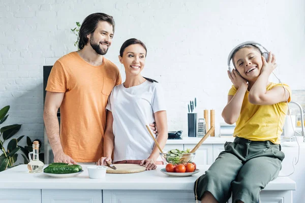 Lächelndes kleines Mädchen mit Kopfhörern auf dem Tisch sitzend, während ihre Eltern sich im Hintergrund umarmen und sie in der Küche anschauen — Stockfoto