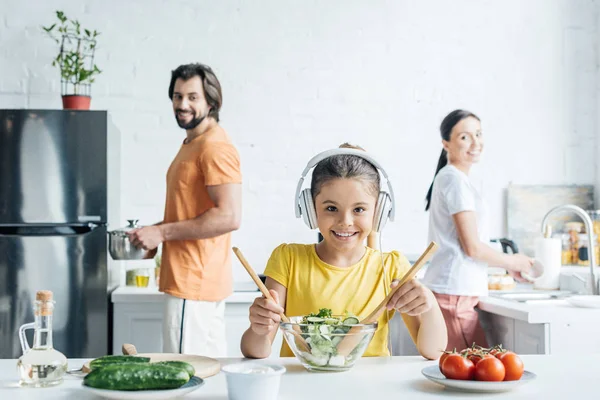 Sorrindo menina em fones de ouvido preparando salada enquanto seus pais de pé borrado no fundo na cozinha — Fotografia de Stock