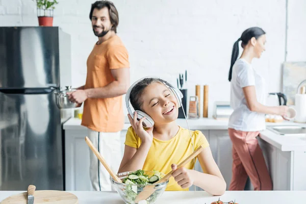 Glückliches kleines Mädchen mit Kopfhörern, das Salat zubereitet, während ihre Eltern in der Küche verschwommen vor dem Hintergrund stehen — Stockfoto