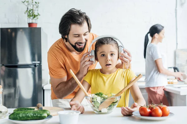 Smiling father and shocked daughter in headphones cooking together while mother washing dishes blurred on background at kitchen — Stock Photo