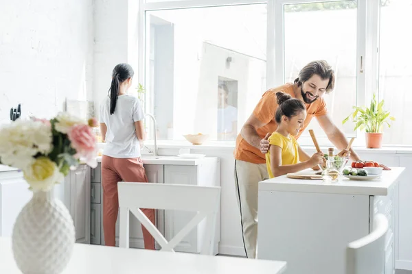 Усміхнений батько і дочка готують разом, а мати миє посуд розмитим на фоні на кухні — стокове фото