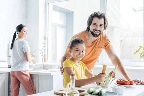 Pai e filha cozinhar juntos, enquanto a mãe lavar pratos borrados no fundo na cozinha — Fotografia de Stock