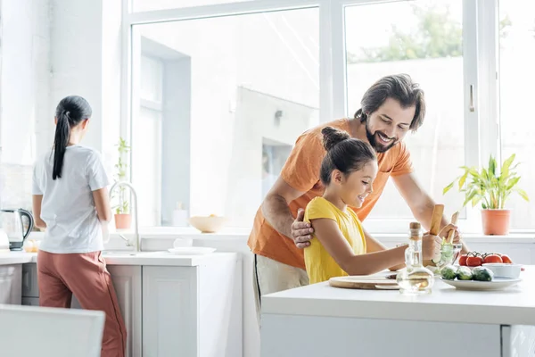 Счастливый отец и дочь готовят вместе, в то время как мать моет посуду размыто на заднем плане на кухне — стоковое фото