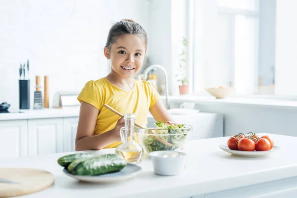 Lächelndes kleines Mädchen, das in der Küche Salat zubereitet und in die Kamera schaut — Stockfoto