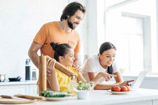 Hermosa familia joven usando el ordenador portátil mientras cocina en la cocina - foto de stock