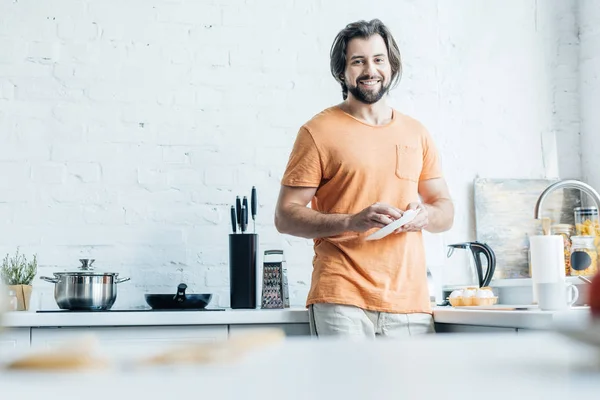Красивый бородатый мужчина моет посуду в одиночестве на кухне и смотрит в камеру — стоковое фото