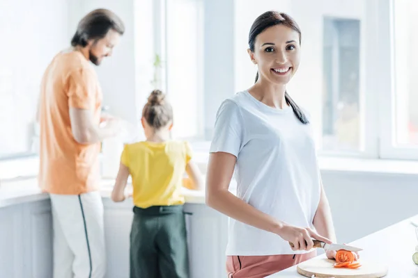 Beautiful young woman cutting tomato for salad while her daughter and husband standing blurred on background — Stock Photo