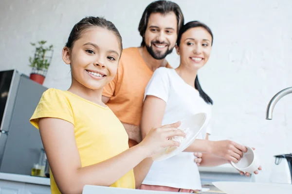 Hermosa familia joven lavar los platos juntos en la cocina y mirando a la cámara - foto de stock