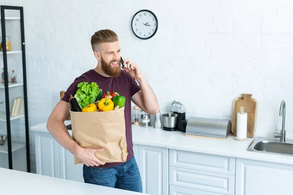 Vista de ángulo alto del hombre barbudo sonriente hablando por teléfono inteligente y sosteniendo bolsa de papel con verduras frescas - foto de stock