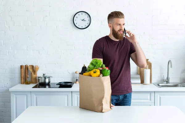 Handsome bearded young man talking by smartphone and looking away in kitchen — Stock Photo