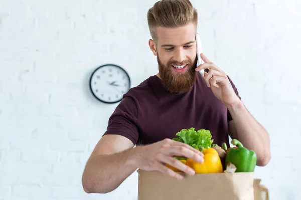 Bel giovane sorridente che parla con lo smartphone e guarda la borsa della spesa con le verdure — Foto stock