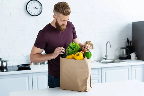 Guapo barbudo joven mirando en la bolsa de comestibles con verduras frescas - foto de stock
