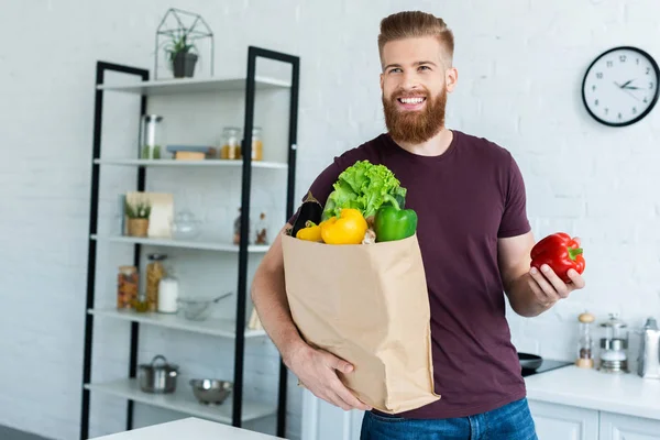 Bel giovane sorridente che tiene la borsa della spesa con verdure fresche in cucina — Foto stock