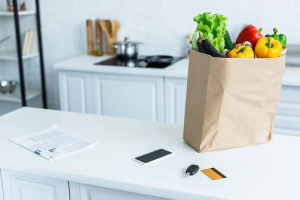 Bolsa de comestibles, smartphone, periódico, llave y tarjeta de crédito en la mesa de la cocina - foto de stock