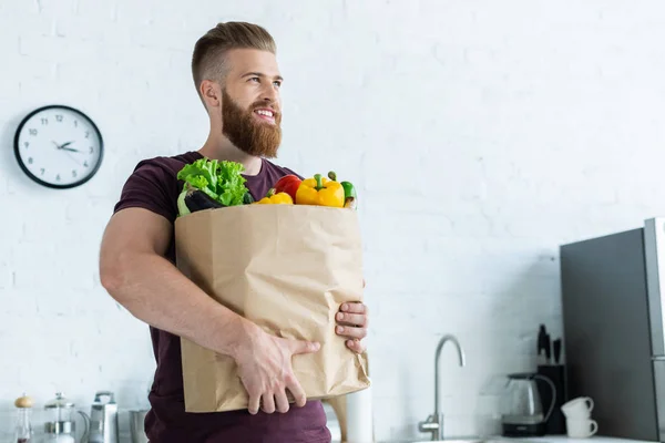 Guapo sonriente joven sosteniendo bolsa de comestibles con verduras y mirando hacia otro lado - foto de stock