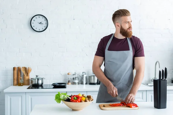 Beau jeune homme barbu dans tablier regardant loin tout en cuisinant dans la cuisine — Photo de stock