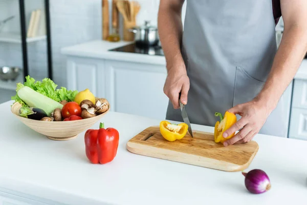 Cropped shot of man in apron cutting bell pepper on wooden cutting board — Stock Photo