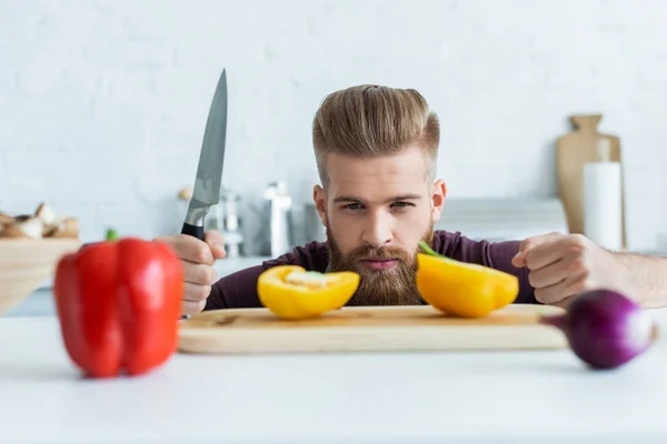 Schöner bärtiger junger Mann mit Messer und Blick auf Paprika — Stockfoto