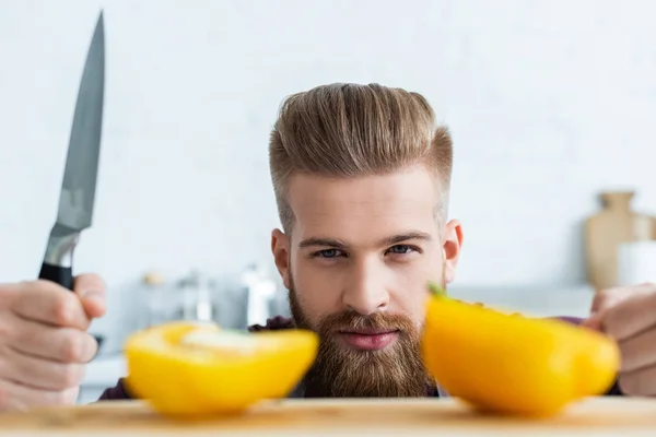 Handsome bearded young man holding knife and looking at halves of bell pepper — Stock Photo