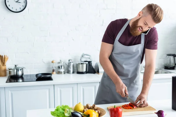 Lächelnder bärtiger junger Mann in Schürze schneidet Gemüse und spricht mit dem Smartphone in der Küche — Stockfoto