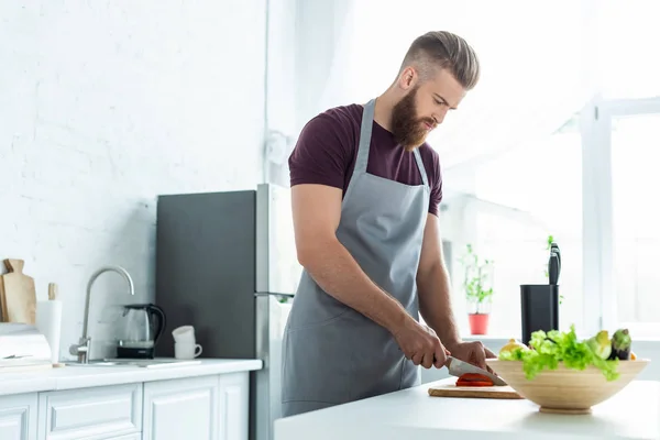 Jovem barbudo bonito em avental cortar legumes enquanto cozinha — Fotografia de Stock