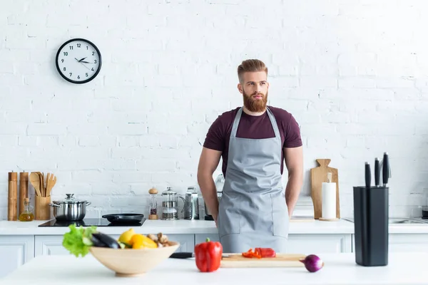 Bello barbuto giovane in grembiule cucina in cucina — Foto stock