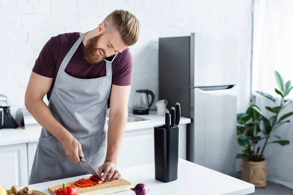 Красивый улыбающийся бородатый мужчина в фартуке разговаривает со смартфоном и режет овощи на кухне — стоковое фото