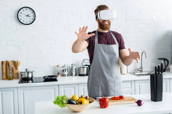 Joven barbudo sonriente en delantal con auriculares de realidad virtual mientras cocina en la cocina - foto de stock