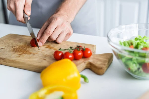 Colpo ritagliato di uomo in grembiule taglio pomodoro ciliegia durante la cottura insalata — Foto stock