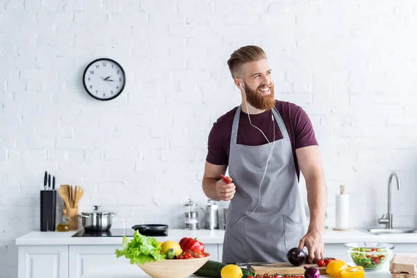 Handsome smiling bearded man in apron listening music in earphones and looking away while cooking in kitchen — Stock Photo